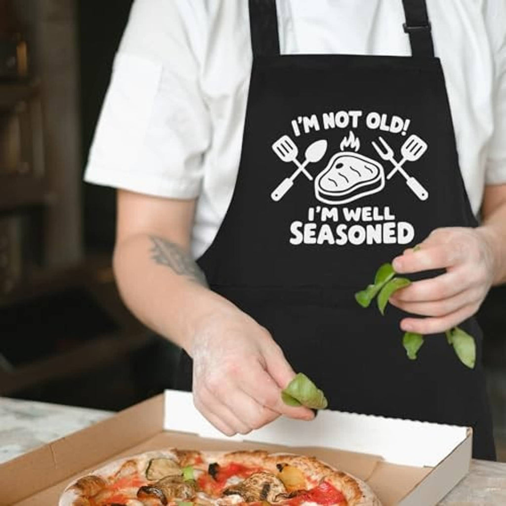 Black apron with "I'm well seasoned" text and utensil graphic while placing food