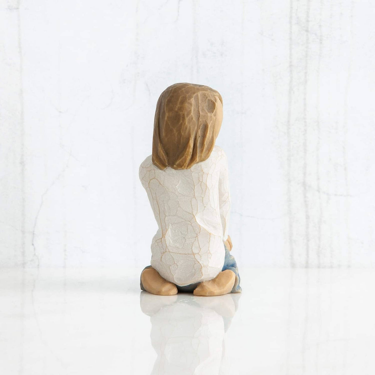 Seated child figurine from behind, brown hair and white robe with blue-gray blanket.