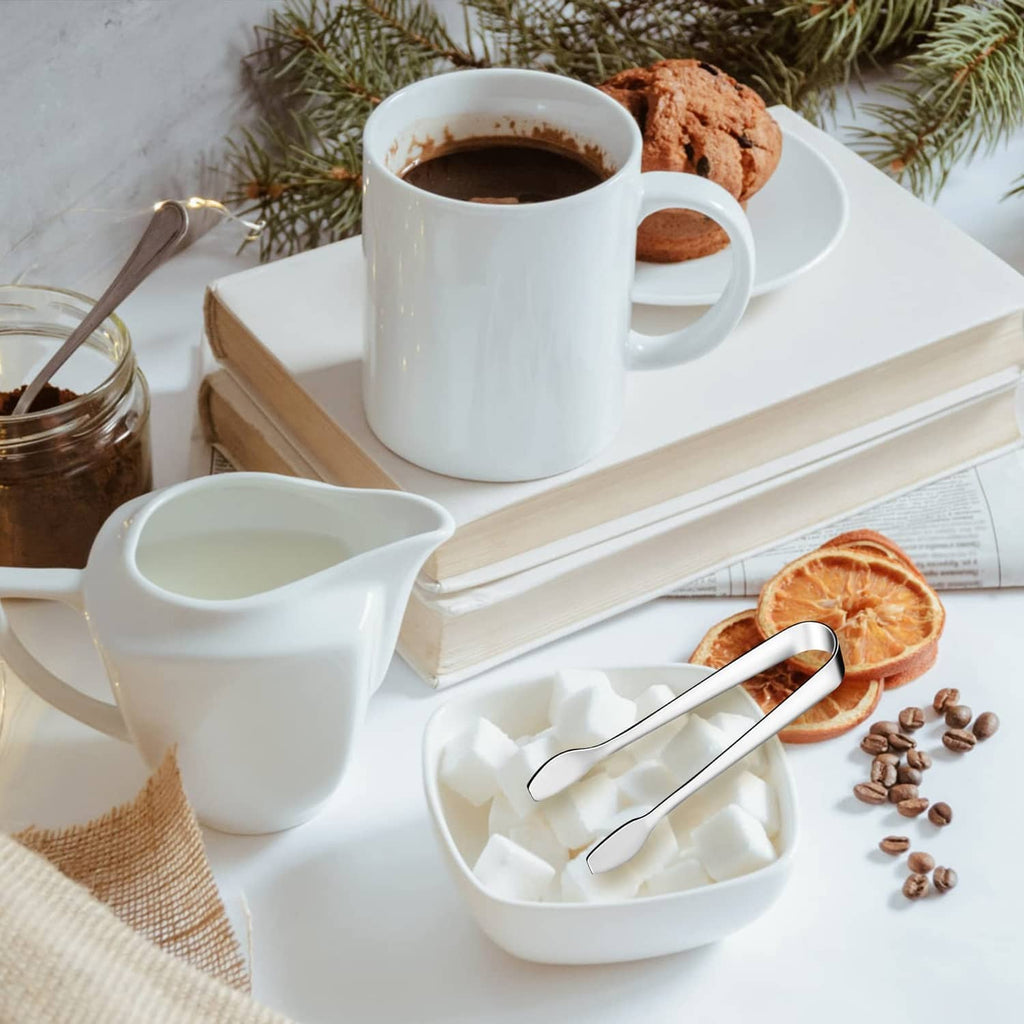 Mini stainless steel serving tongs on a tray beside a cup and small plates