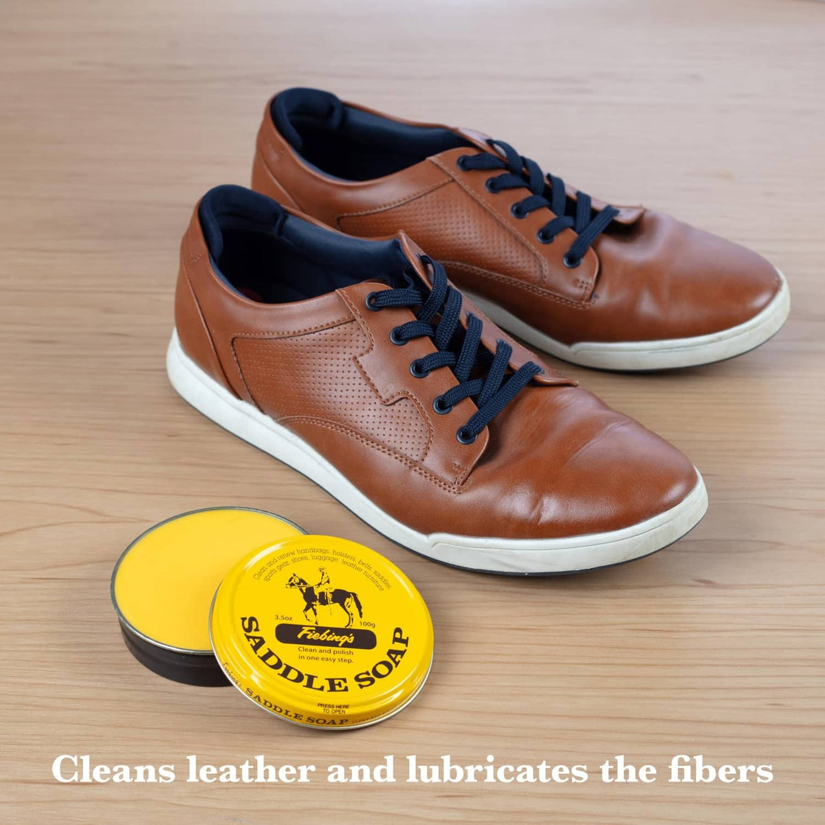 Yellow saddle soap tin next to brown leather sneakers on a light wood surface