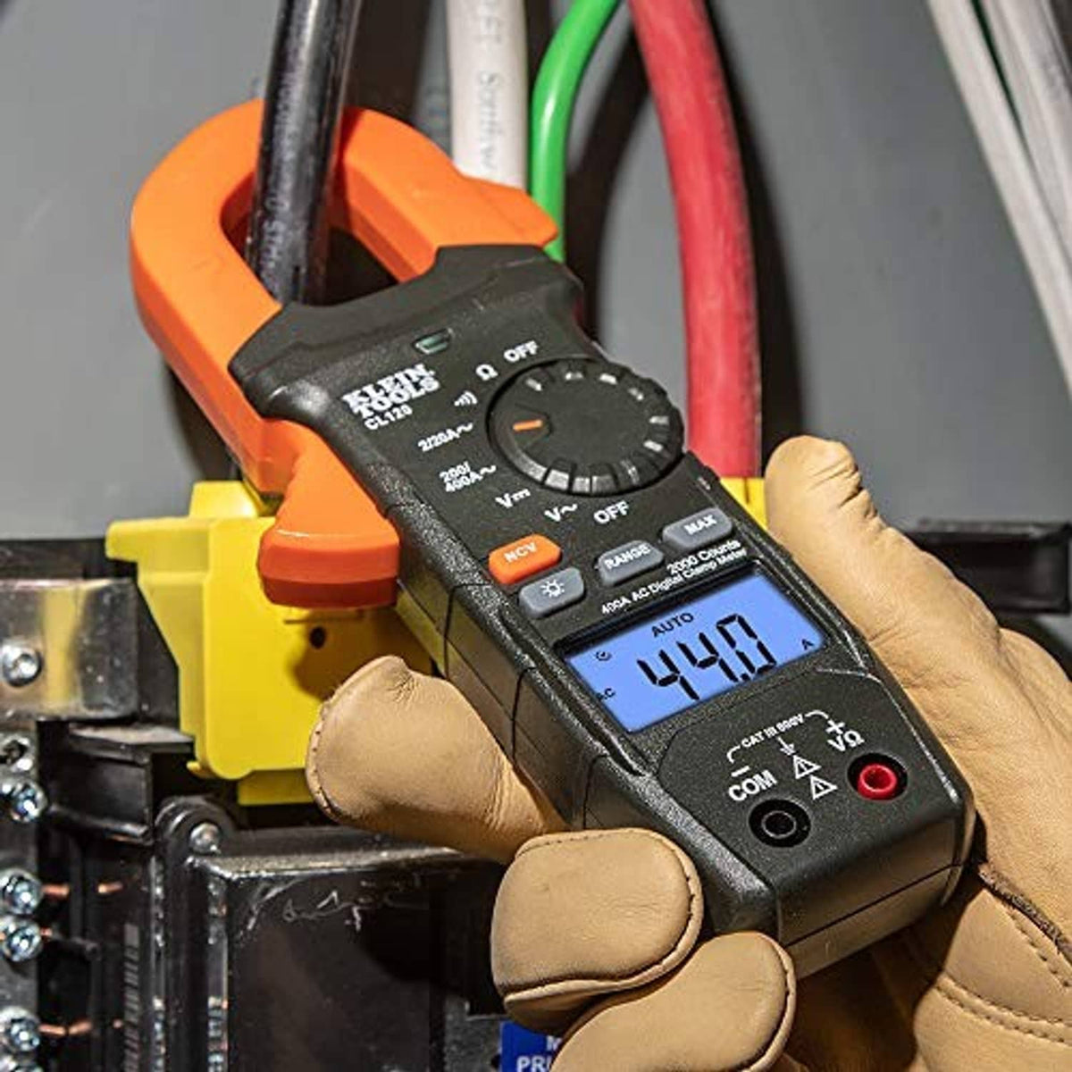 Digital clamp meter with orange clamp and blue display, held by a gloved hand.