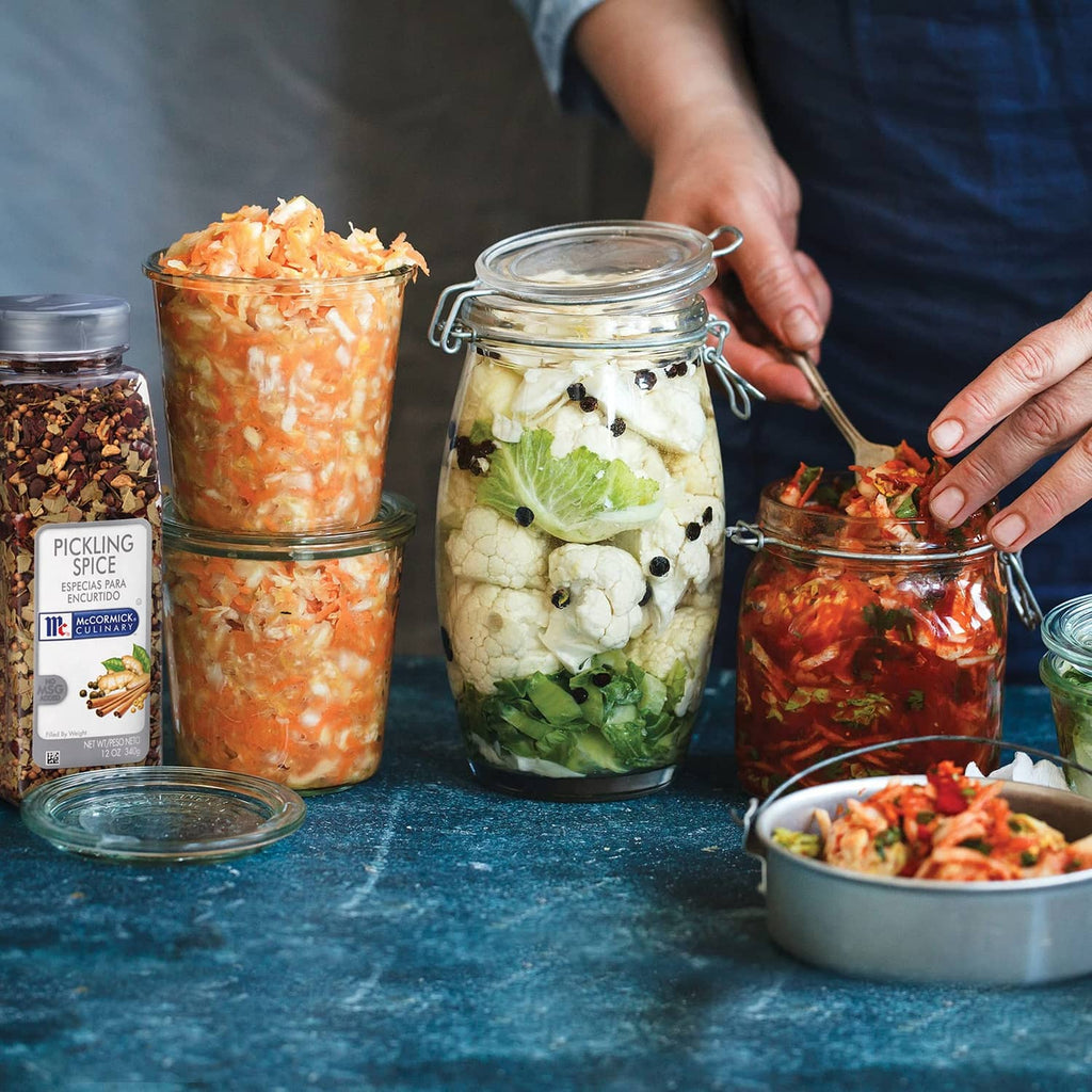 Person arranging glass jars of pickled vegetables beside containers of pickling spice