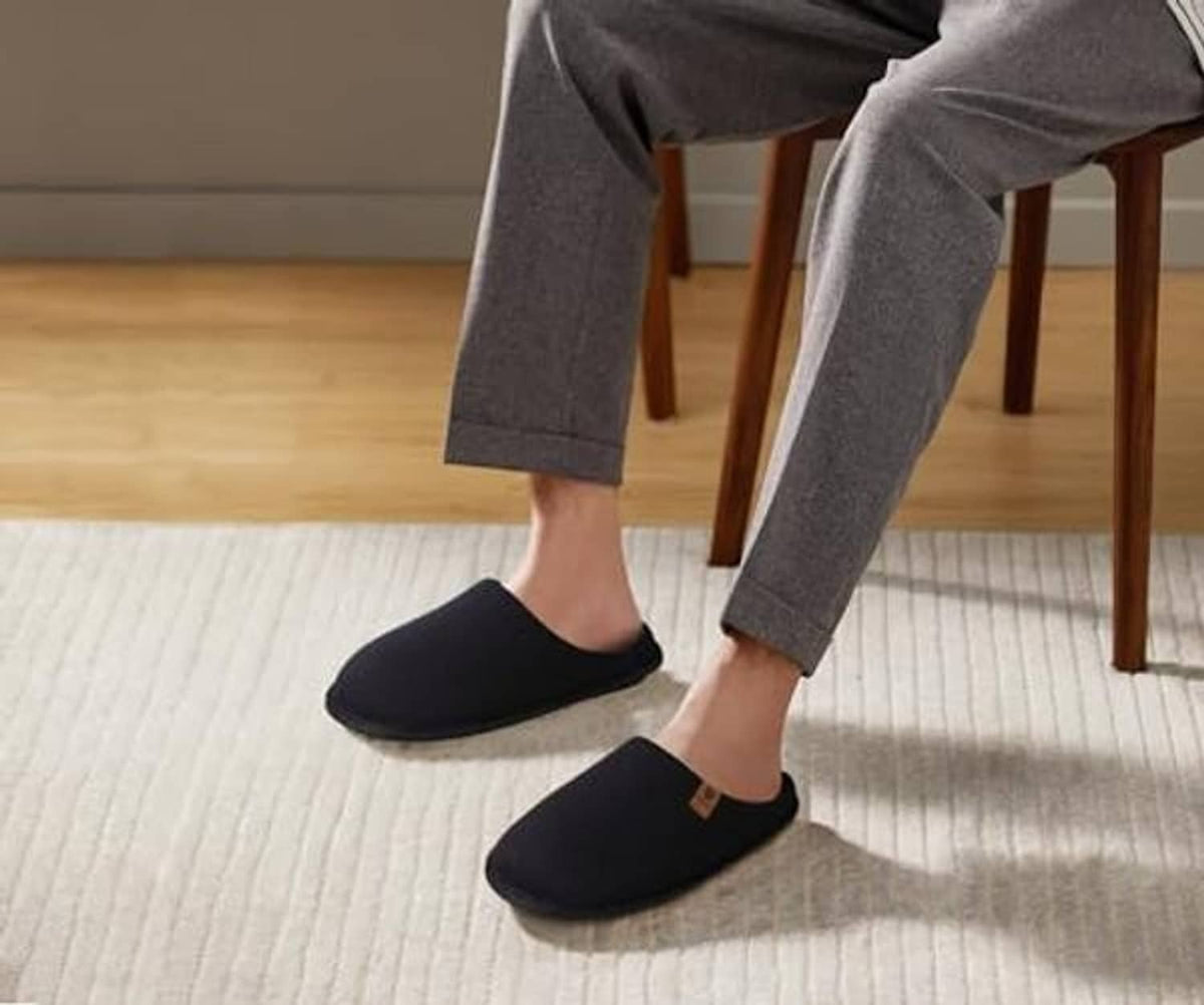 Pair of men's black slip-on house slippers on a light indoor rug.