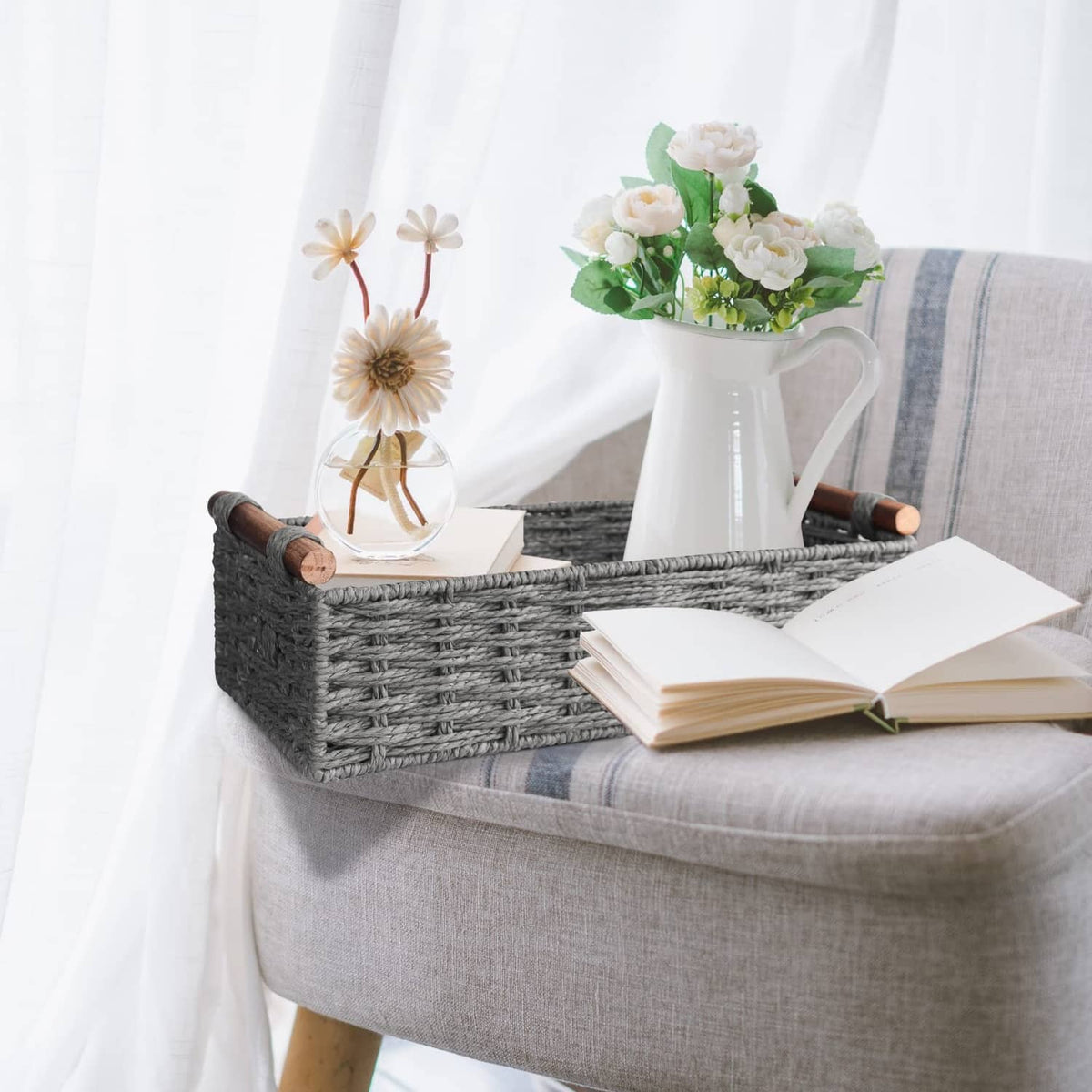 Grey woven storage basket with wooden handles on a couch; open book and flowers inside.