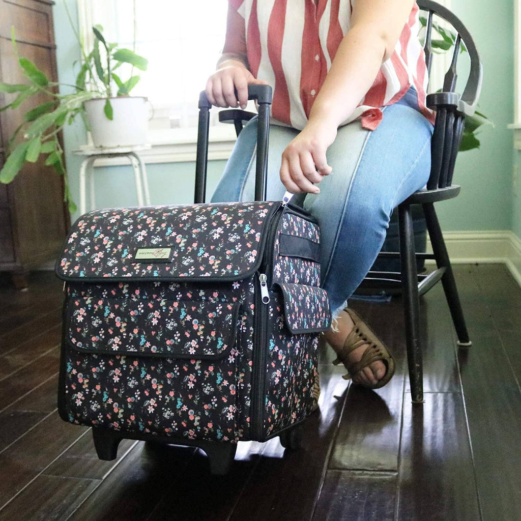 Black floral rolling machine tote on wheels with front pocket and flap, held by person