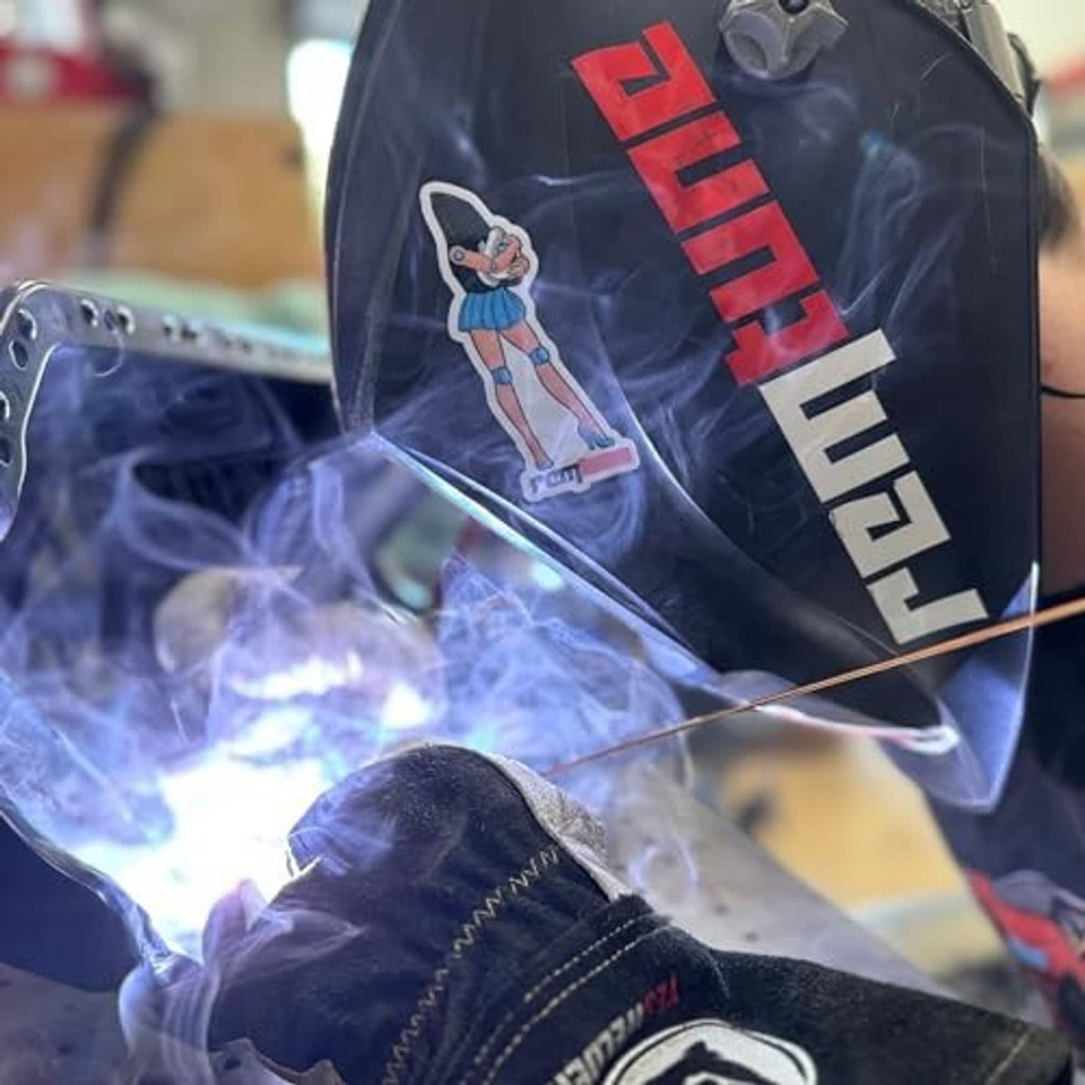 Weld-on bung fitting being welded to metal plate with visible torch sparks and smoke