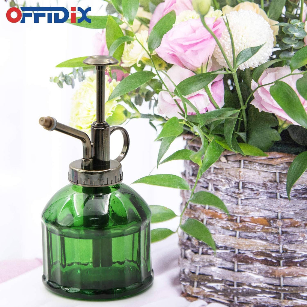 Green glass plant mister with bronze sprayer top and curved spout, near garden decor.