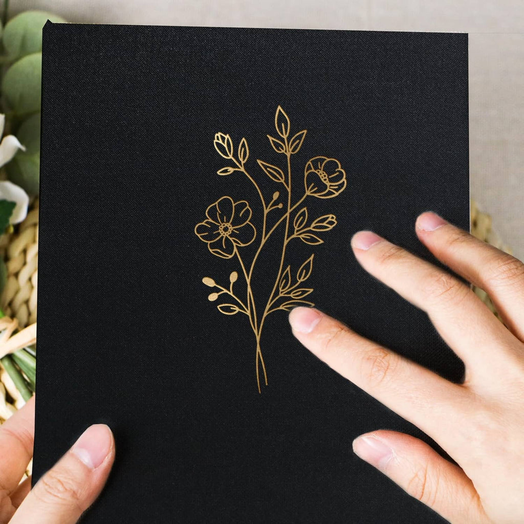 Black photo album front cover with gold floral tree line art, held by hands