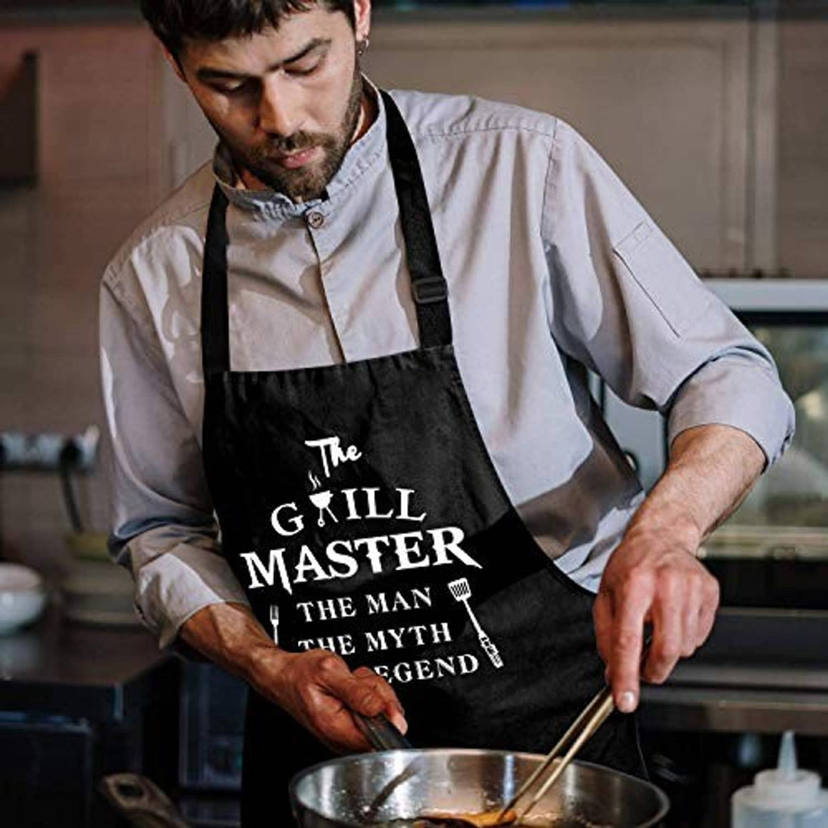 Man wearing a black grill-themed apron over a white shirt in a kitchen.
