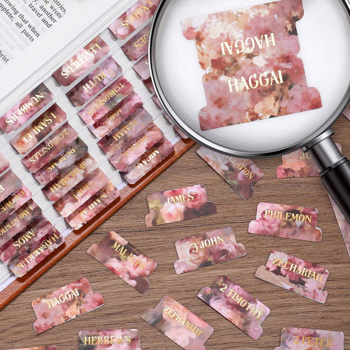 Pink floral Bible index tabs with gold-foil book names, some loose on a wooden surface.