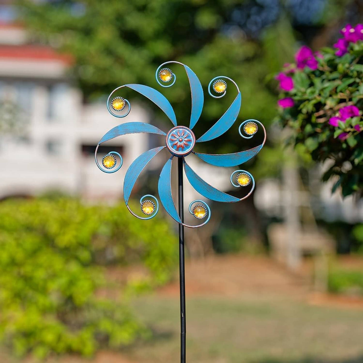 Outdoor blue metal wind spinner with curved blades, jeweled hub, and gold accent rings on pole.