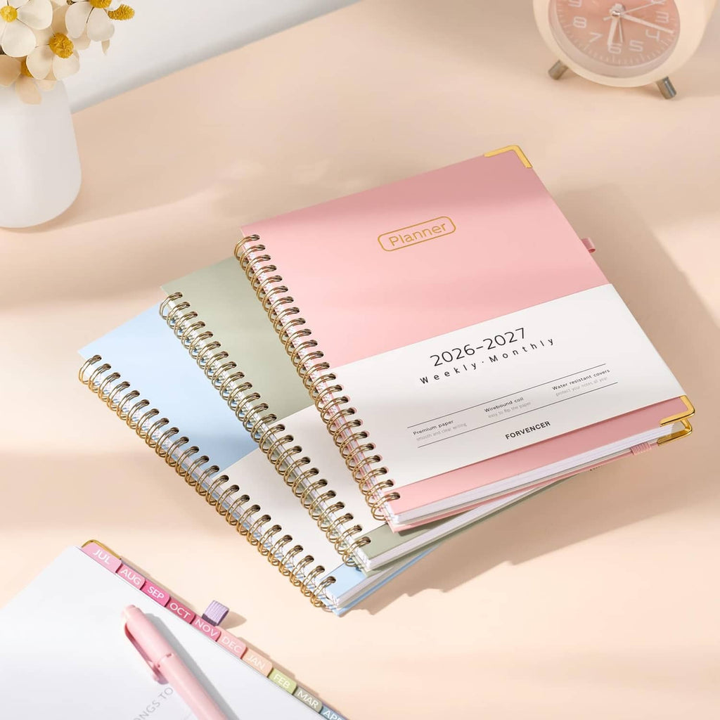 Pink spiral planner stack with gold ribbon bookmark; dated 2026–2027 weekly monthly cover