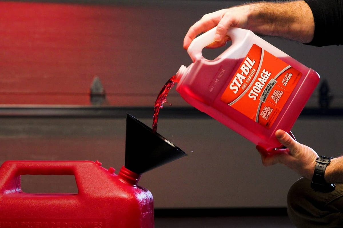 Hands pouring red fuel stabilizer from white spout jug into red fuel container