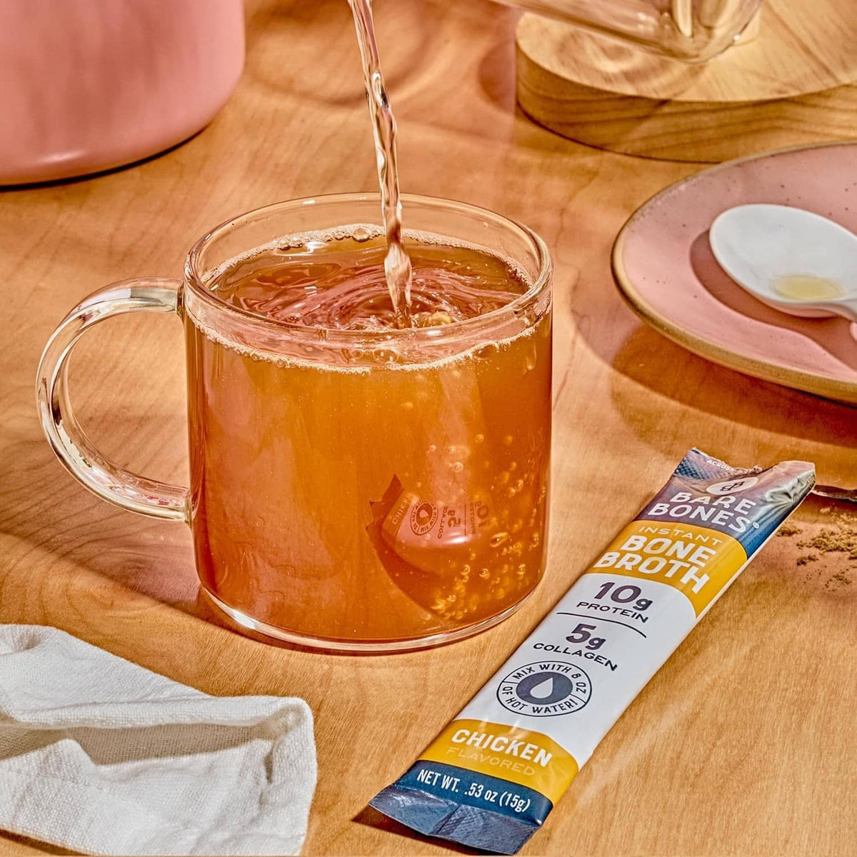 Instant bone broth powder stick pack next to a glass mug with hot broth pour
