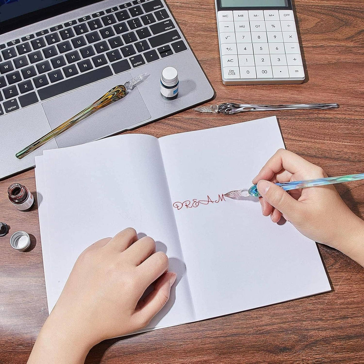 Glass dip pen held while writing in an open notebook on a wooden desk with ink bottle