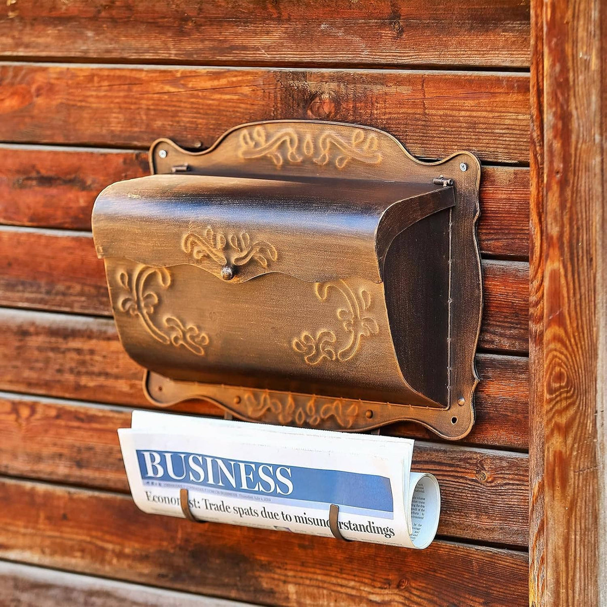 Dark bronze wall mailbox with embossed design and newspaper holder filled with documents