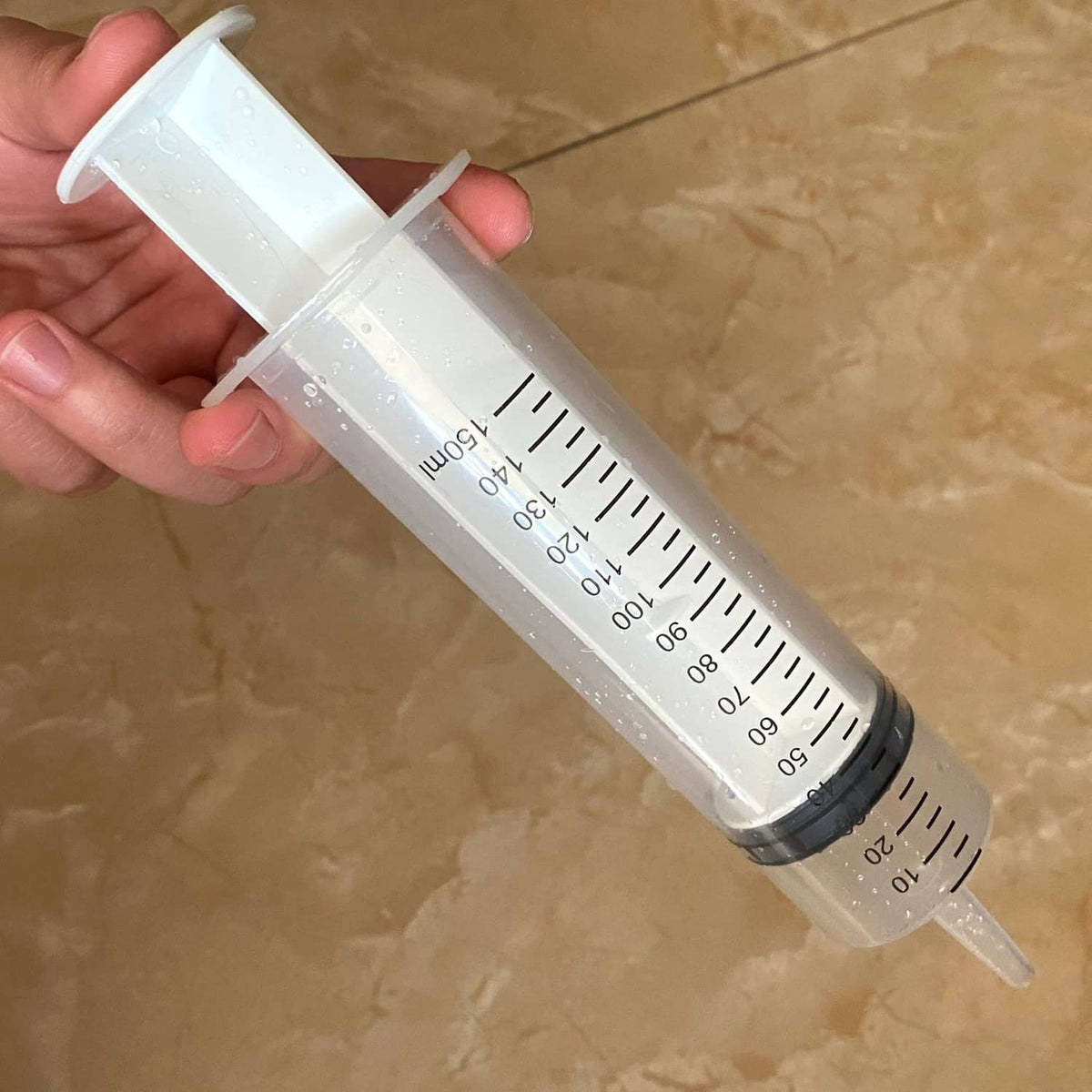 Hand holding clear 150 ml syringe with black measurement scale and gray plunger