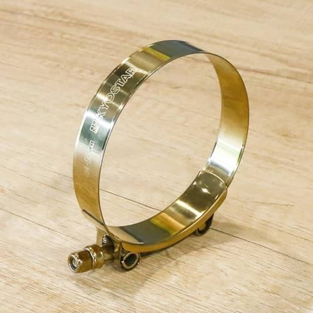 Gold stainless T-bolt hose clamp resting on wood surface, side screw visible.