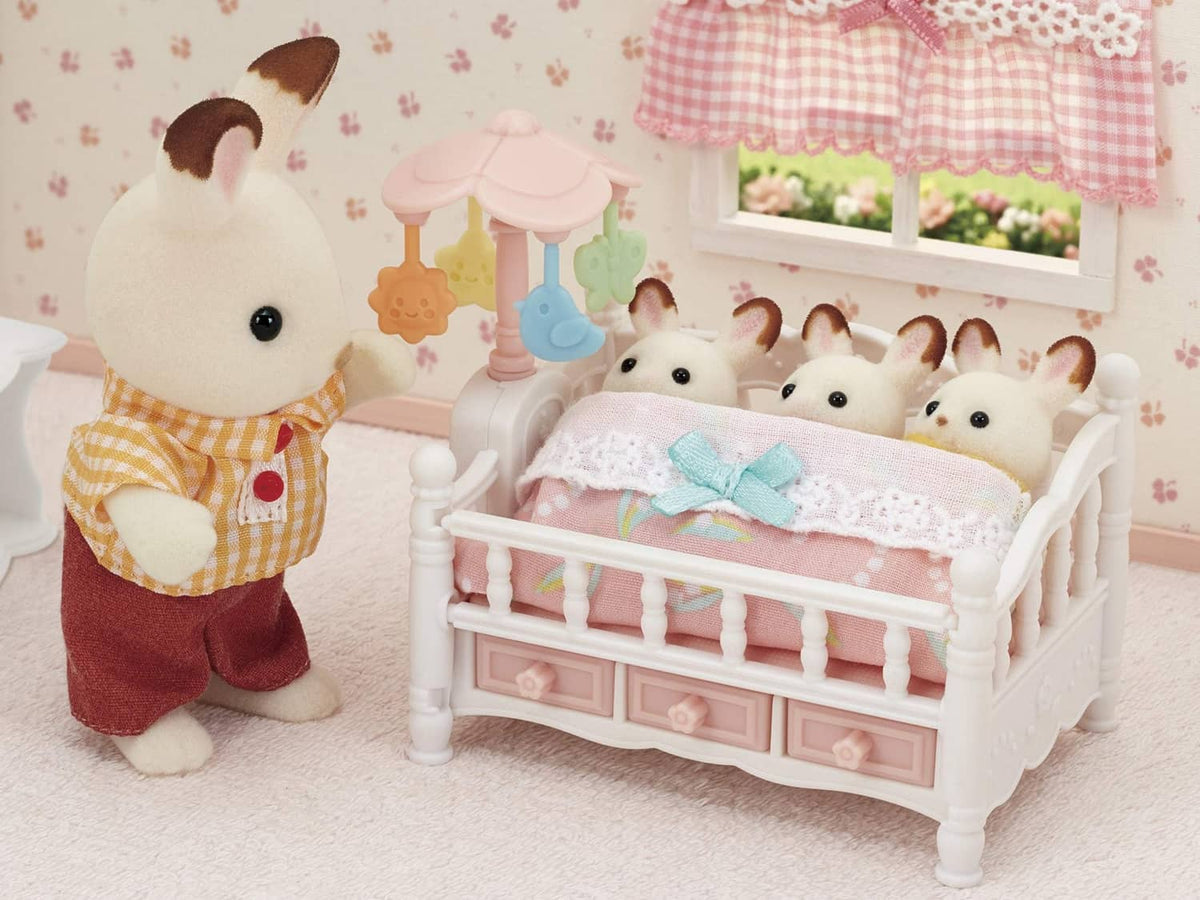 White dollhouse crib with pink blanket and blue bow, with bunny dolls and mobile stand