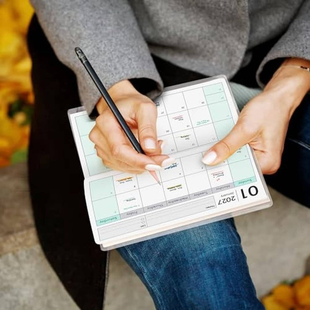 Pocket calendar planner held in hands with a pen writing on the monthly grid