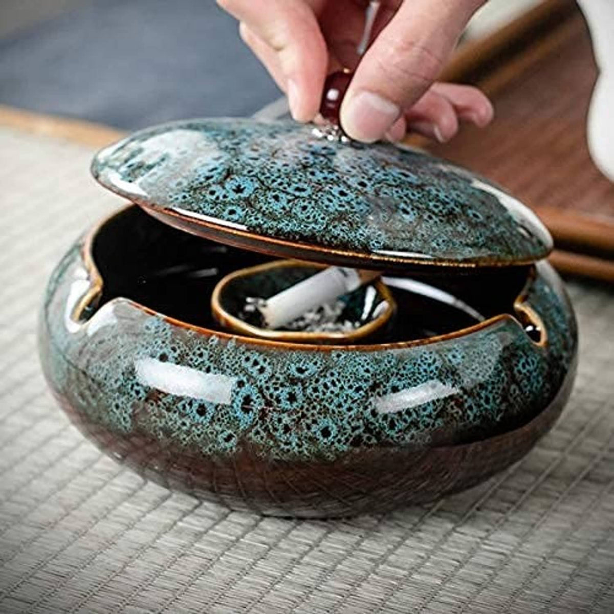 Open turquoise ceramic ashtray with hinged lid and brown interior, hand lifting lid