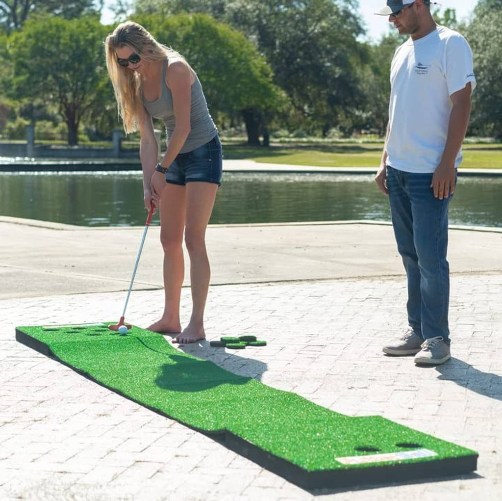 Green turf putting mat on pavement with multiple holes and a red putter.