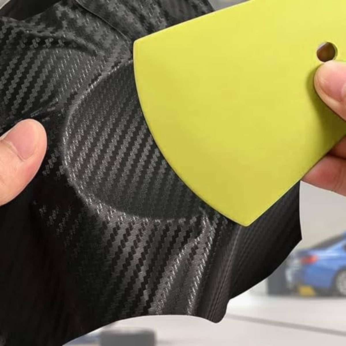 Yellow squeegee tool smoothing vinyl on a dark carbon-fiber style surface