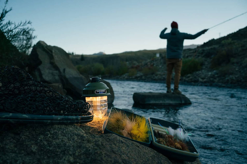 Olive drab camping lantern with warm LED glow on rock beside folded gear
