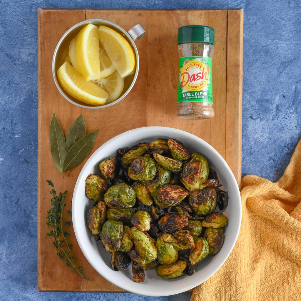 Salt-free table seasoning blend bottle with lemon halves, herbs, and roasted Brussels sprouts.