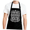 Black apron with adjustable neck strap and large vintage-style front logo patch.