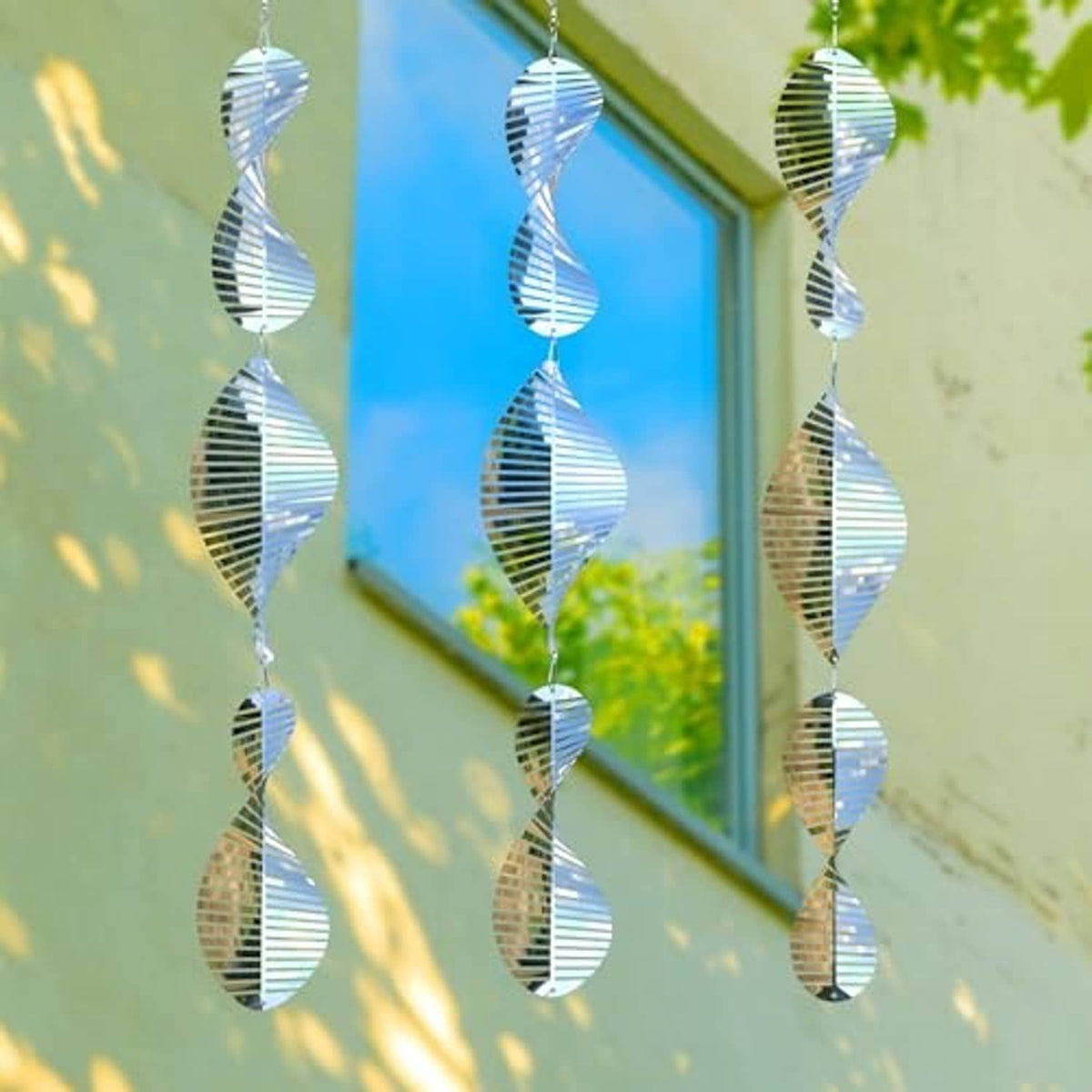 Stainless steel bird-scarer wind spinners hanging near a window, leaf-dappled light