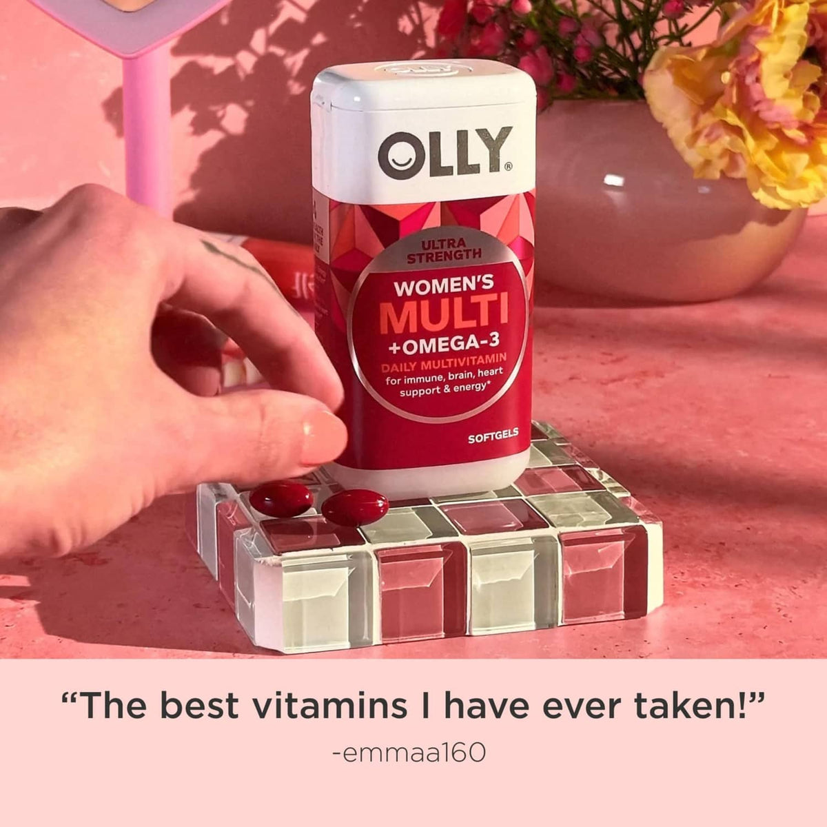 Hand holding OLLY Women's Multi +Omega-3 softgels bottle with red capsules.