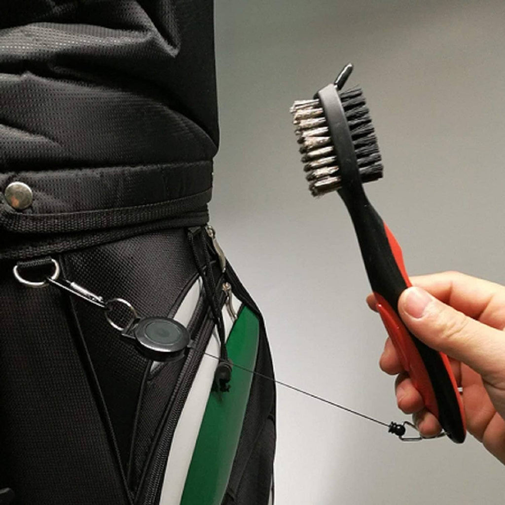 Golf club groove cleaner brush with red-and-black handle, held in hand near a golf bag