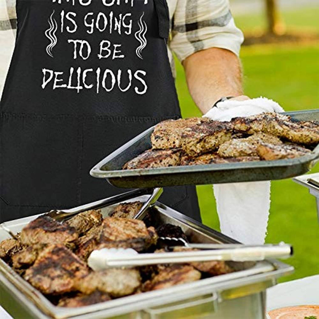 Black BBQ apron with funny slogan beside grilled meat tray and tongs, outdoor cooking scene