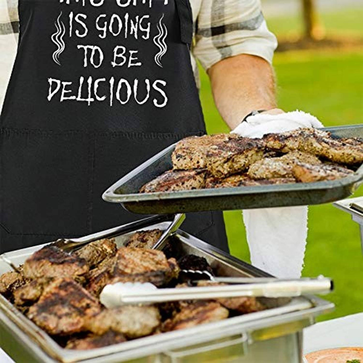 Black BBQ apron with funny slogan beside grilled meat tray and tongs, outdoor cooking scene