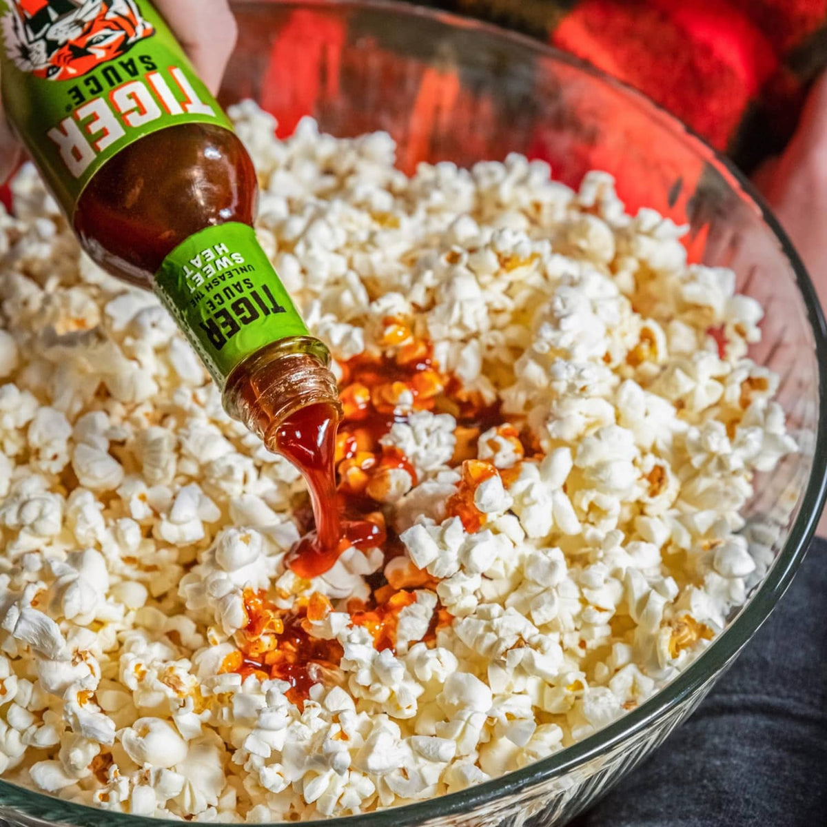 Hot sauce bottle pouring dark red sauce over popcorn in a glass bowl