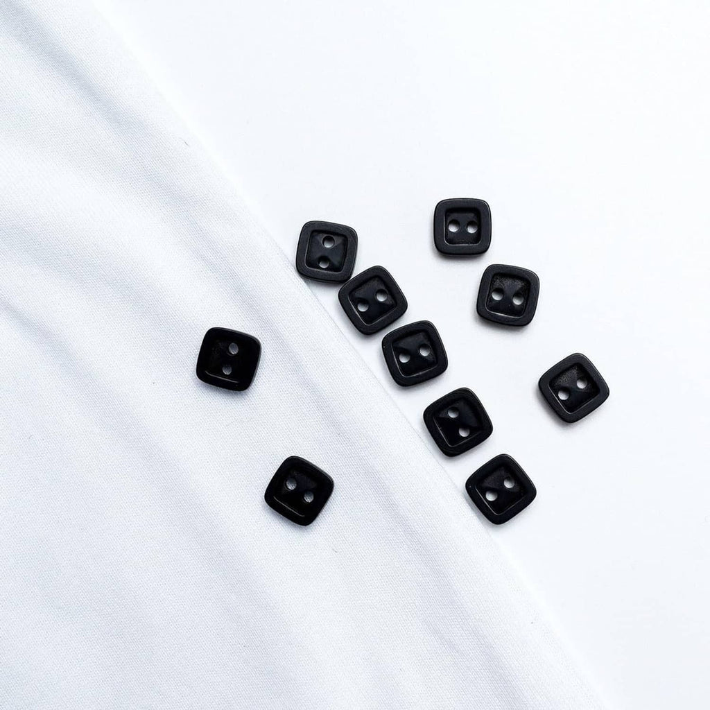 Black square 2-hole resin buttons scattered on white fabric, multiple front designs