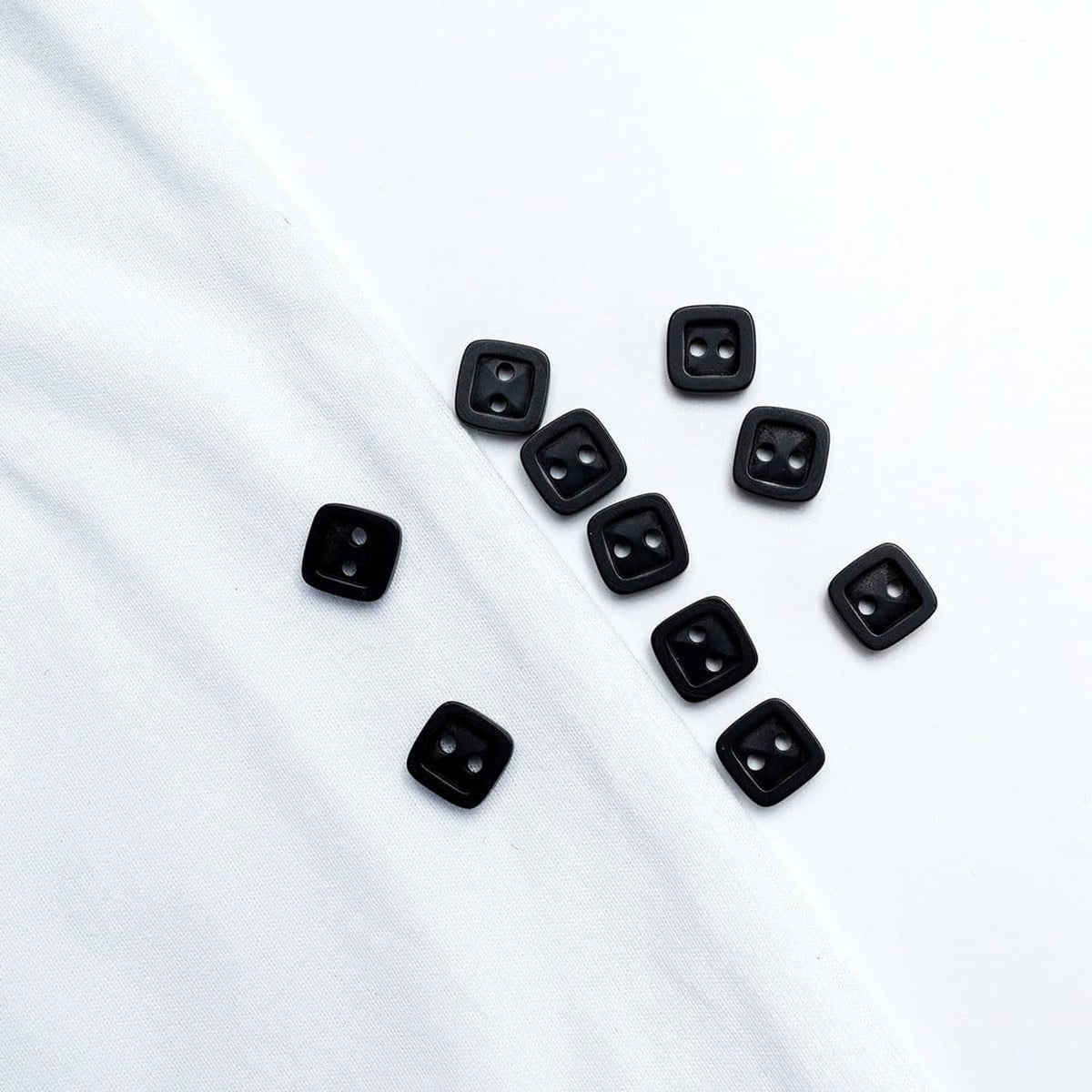 Black square 2-hole resin buttons scattered on white fabric, multiple front designs