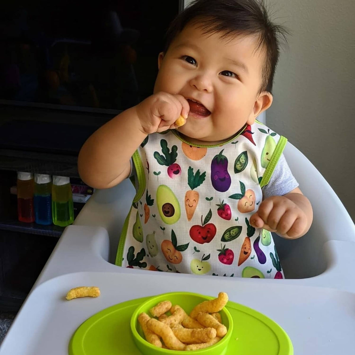 Baby in high chair eating puff snacks from a bowl and holding a piece