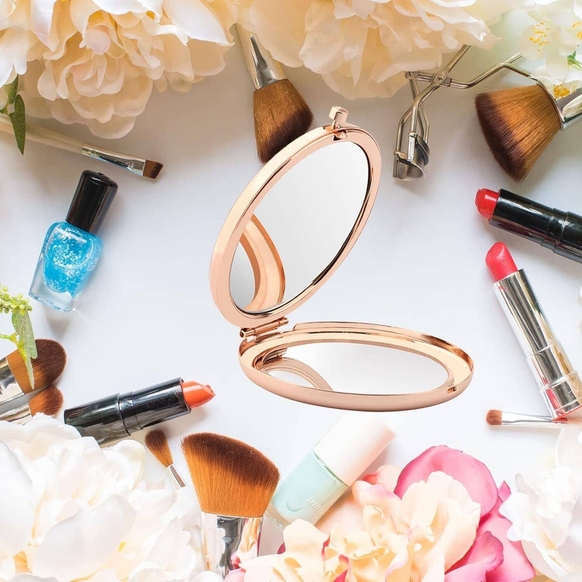 Rose-gold oval makeup mirror compact with flip-open lid, surrounded by cosmetics.