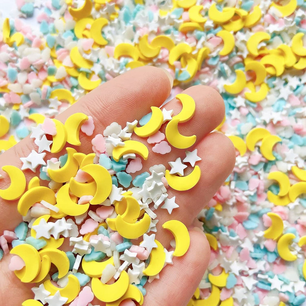 Hand holding small resin sprinkles: yellow moons, pastel clouds, and white stars