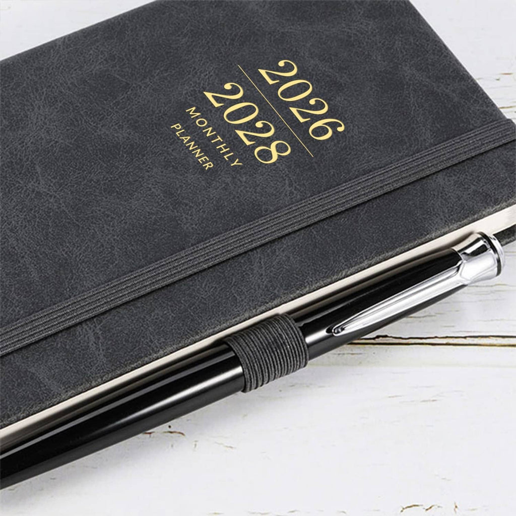 Dark faux-leather pocket planner cover with gold “2026–2028” text and pen on desk
