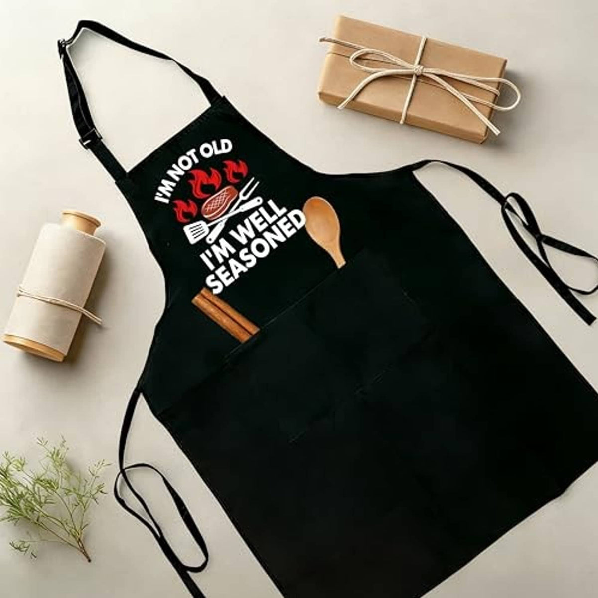 Black apron laid flat with printed “I’m well seasoned” quote and utensil graphics