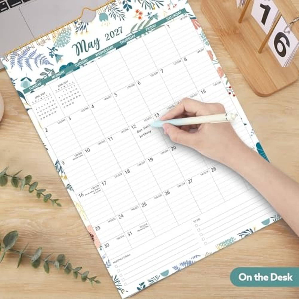 Floral wall calendar showing May 2027, with daily grid and a hand writing in pencil