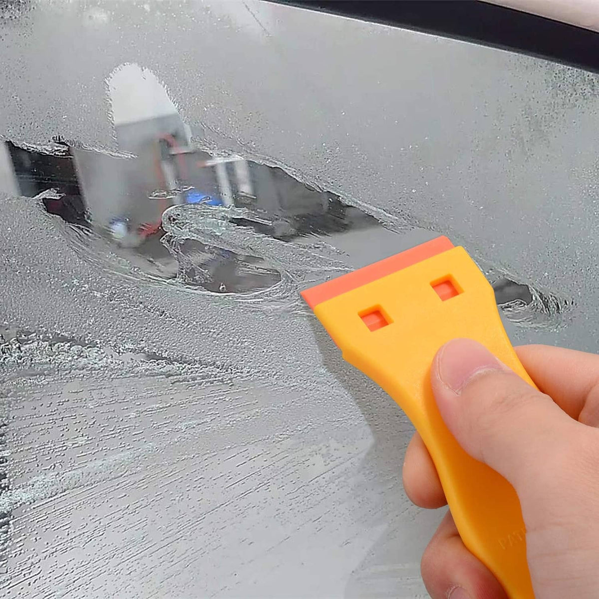 Handheld yellow plastic scraper removing film from metal surface, orange blade base