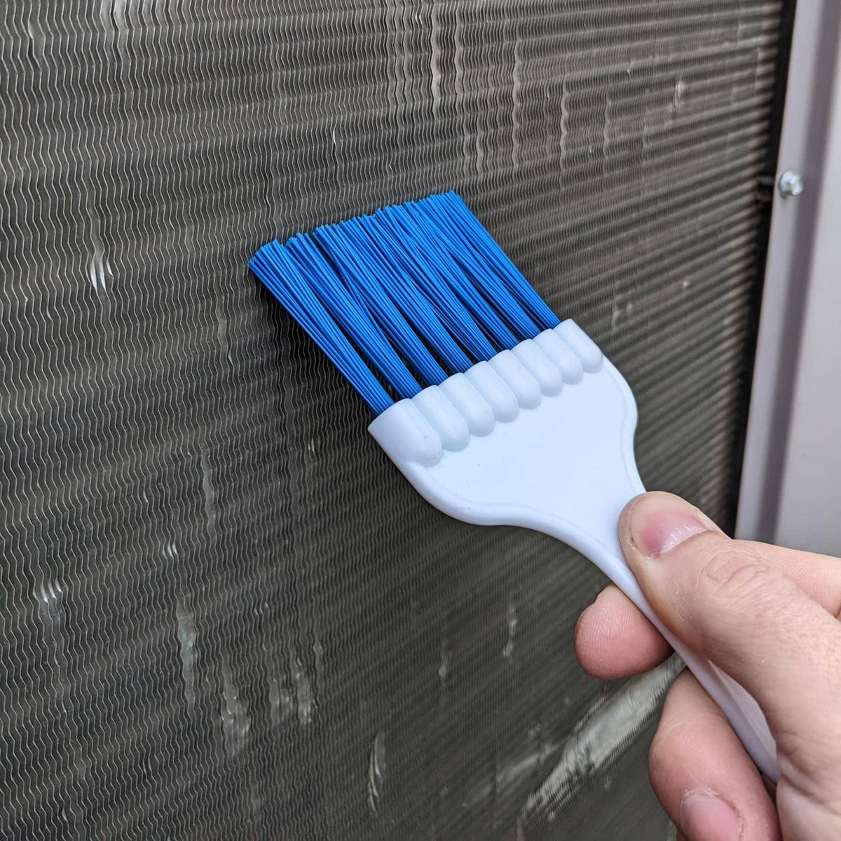 Handheld coil cleaning brush with blue bristles scrubbing a metal grille