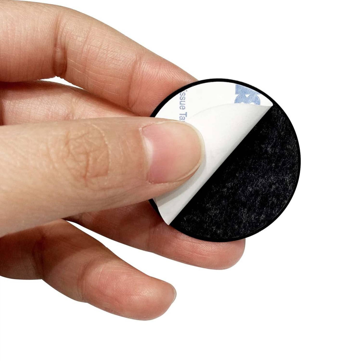 Hand peels backing from black adhesive metal plate disk, exposing sticky adhesive