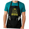 Black cooking apron on teal shirt with adjustable straps and utensil graphic