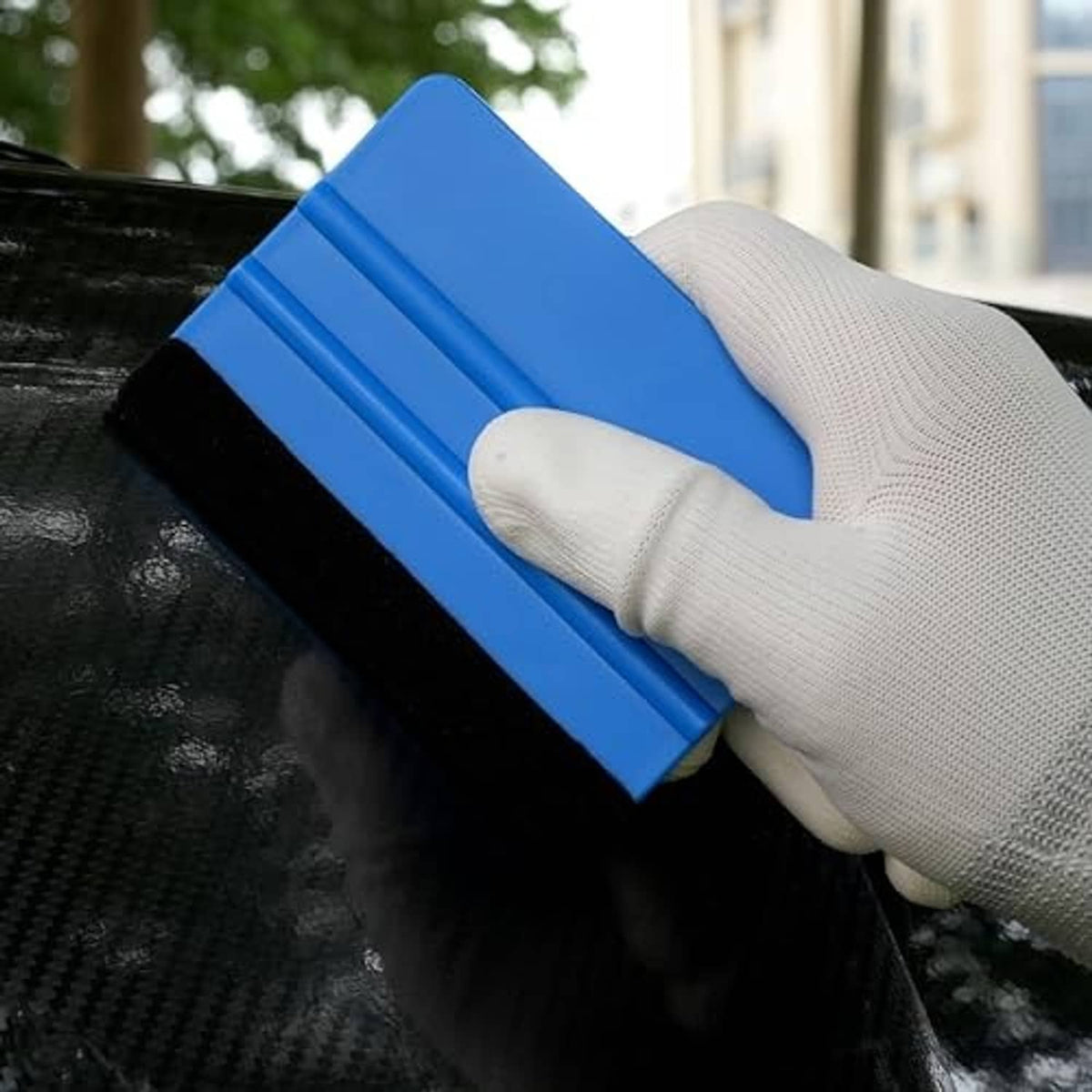 Blue squeegee tool with black felt edge, held by gloved hand on dark surface