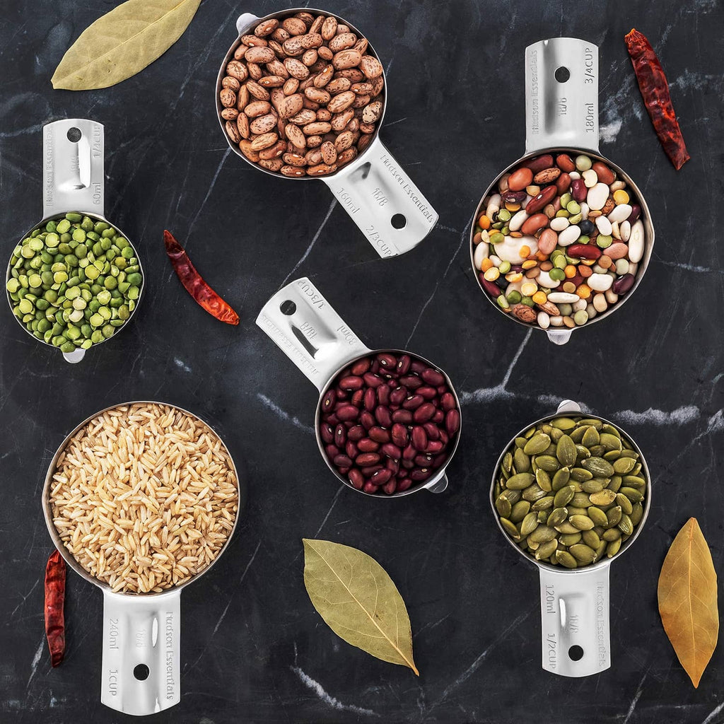 Six stainless steel measuring cups filled with assorted beans and seeds